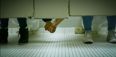 Two people in separate bathroom stalls reaching under the stalls and holding hands in support